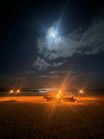 Night beach bonfire on a sandy shore with tiki torches and people gathered around a glowing fire, ocean waves reflecting a bright full moon and cloud halo