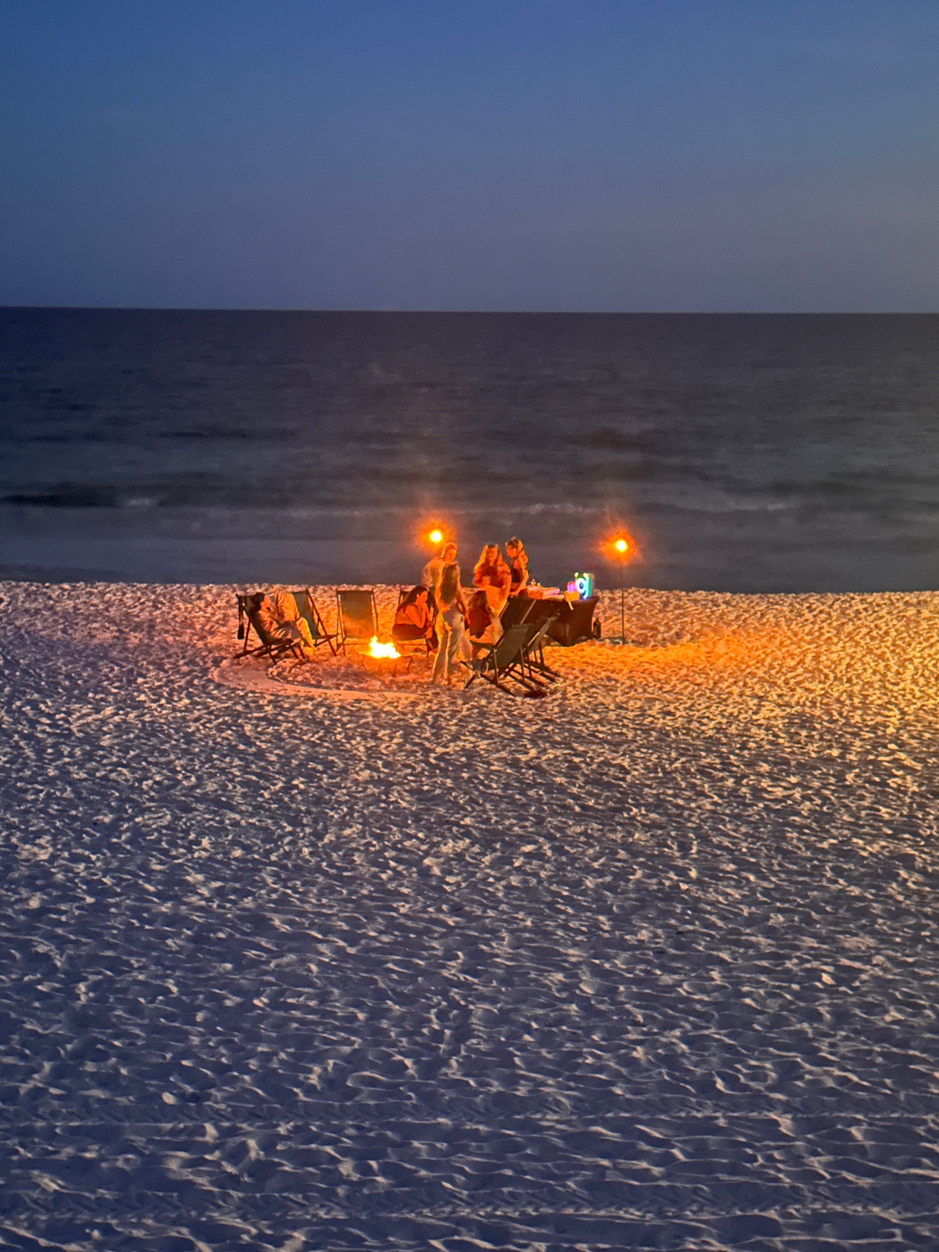 Cozy group gathered around a glowing bonfire and tiki torches on a sandy beach at dusk, folding chairs lit against the dark ocean horizon.
