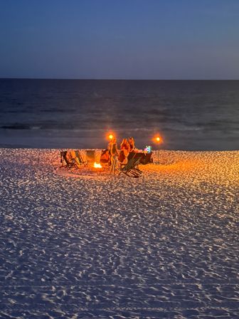 Cozy group gathered around a glowing bonfire and tiki torches on a sandy beach at dusk, folding chairs lit against the dark ocean horizon.
