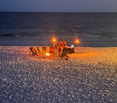 Cozy group gathered around a glowing bonfire and tiki torches on a sandy beach at dusk, folding chairs lit against the dark ocean horizon.