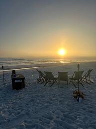 Golden sunset over the ocean with calm waves on a white-sand beach, a row of empty folding chairs and tiki torches beside a small pile of firewood — peaceful seaside evening