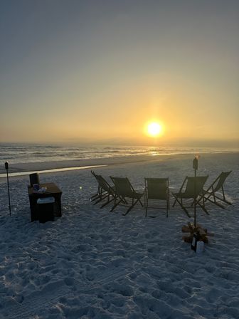 Golden sunset over the ocean with calm waves on a white-sand beach, a row of empty folding chairs and tiki torches beside a small pile of firewood — peaceful seaside evening