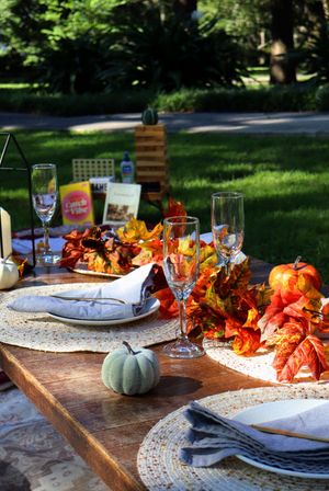 Outdoor fall tablescape on a wooden table in a park featuring woven placemats, folded gray napkins, empty champagne flutes, small decorative pumpkins and bright orange autumn leaf garland on a green lawn.