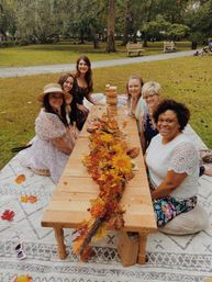 Six women enjoying an autumn picnic in a public park, seated on blankets around a low wooden table with a sunflower and fall foliage centerpiece and a wooden block tower game, smiling under oak trees.