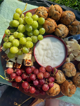 Sunlit picnic snack board with green and red grape clusters, grilled cheese cubes, sliced cheese, blueberry mini muffins and a bowl of creamy yogurt dip