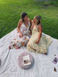 Two friends on a summer picnic blanket on a green lawn, wearing floral sundresses and chatting; round pink cushion with a Savannah travel guide and a pink smartphone nearby.