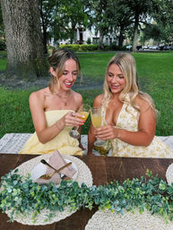 Two women in yellow sundresses clinking mimosa glasses at a rustic picnic table on a green park lawn under large trees, with woven placemats, greenery centerpiece and summer brunch table setting.