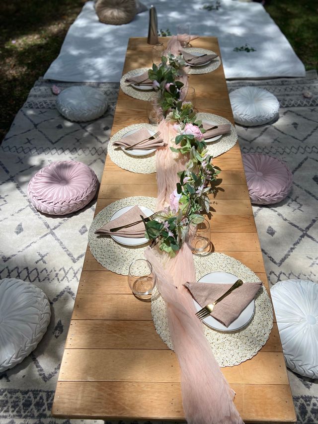 Boho outdoor low wooden picnic table on patterned rugs set for brunch with blush pink chiffon runner, eucalyptus garland, woven placemats, folded napkins, gold flatware and round floor cushions