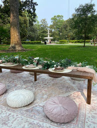 Low garden picnic in a leafy park: patterned rug with round pink and white floor cushions, low wooden table set with eucalyptus garland, plates and glasses, green lawn and decorative fountain in the background.