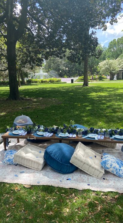 Blue-and-white picnic setup on a blanket under large trees in a sunny park, low wooden table with cushions, floral garland and a fountain visible in the background.