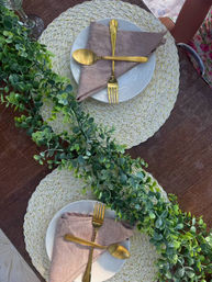 Top-down view of a rustic outdoor table set for two with woven round placemats, white plates, neutral linen napkins, crossed gold flatware and a lush eucalyptus table runner.