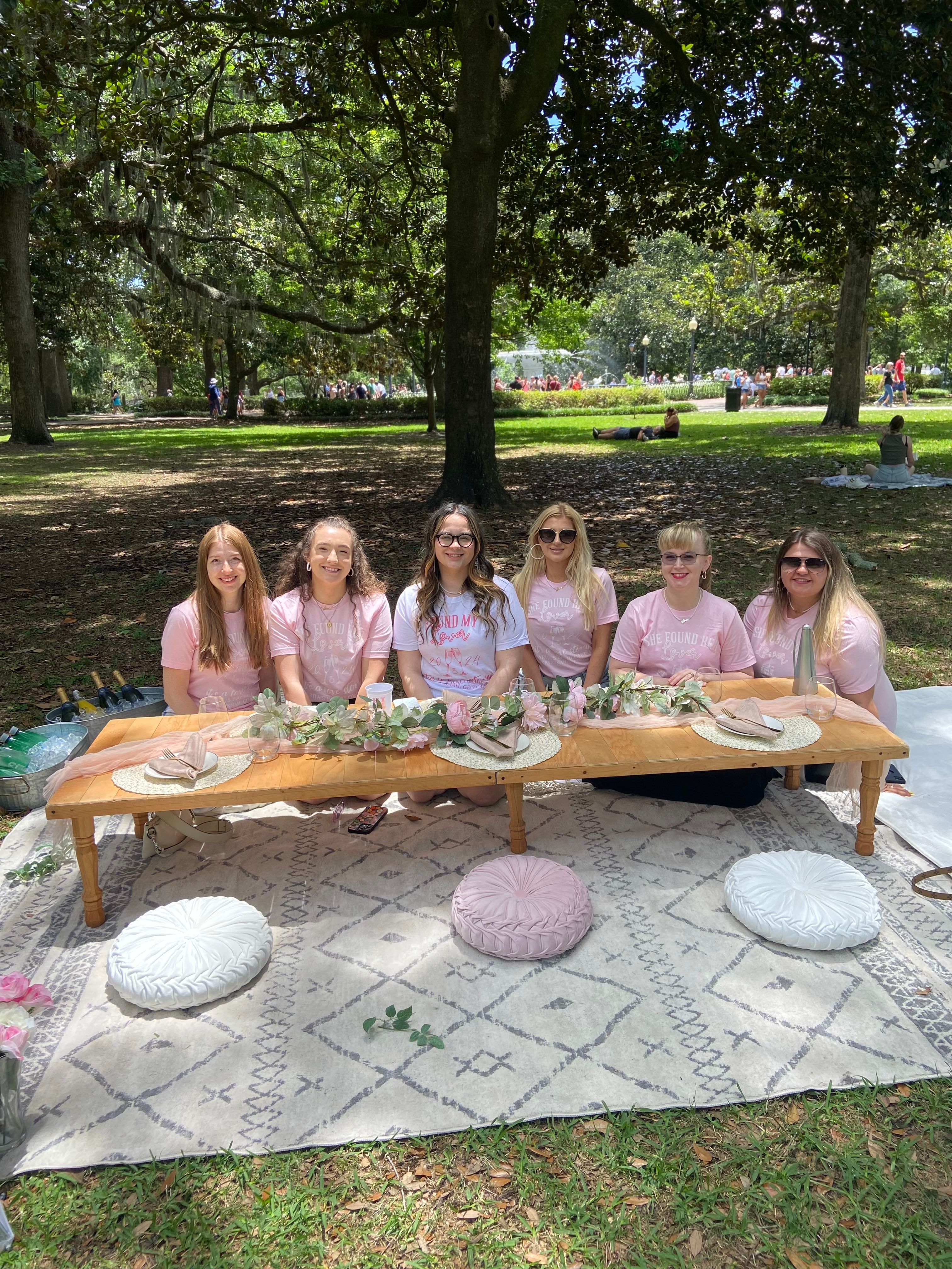 Six women at a pastel-themed outdoor picnic in a shaded city park under large oak trees, seated at a low wooden table decorated with blush floral garland, round cushions on a patterned rug, relaxed sunny gathering.
