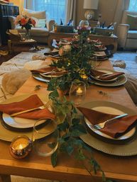 Intimate living-room tablescape on a low wooden table with a greenery runner, gold chargers, rust-colored napkins, glassware and glowing votive candles, ready for guests.