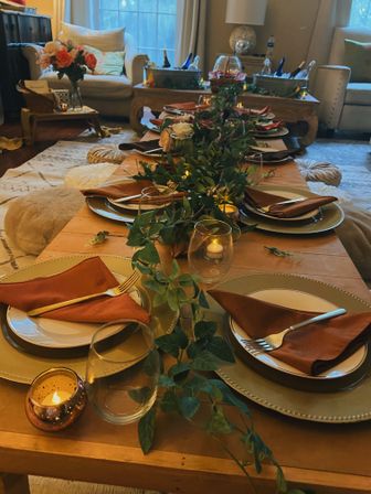 Intimate living-room tablescape on a low wooden table with a greenery runner, gold chargers, rust-colored napkins, glassware and glowing votive candles, ready for guests.