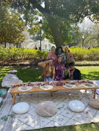 Six friends posing at a boho-style picnic in a sunny city park with a fountain, low wooden table decorated with a floral garland, woven placemats and cushions on a patterned rug under a large tree.