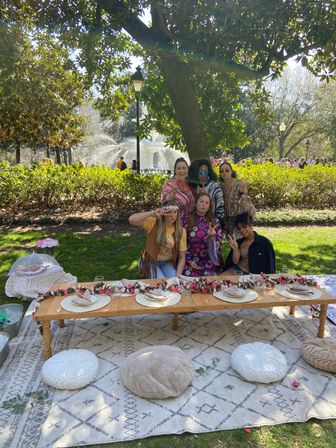 Six friends posing at a boho-style picnic in a sunny city park with a fountain, low wooden table decorated with a floral garland, woven placemats and cushions on a patterned rug under a large tree.