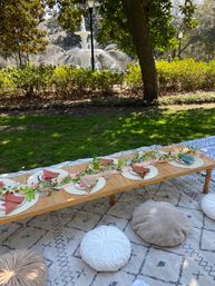 Sunlit park picnic tablescape by a decorative fountain — low wooden table with round placemats, folded napkins, glassware and a greenery garland, plush floor cushions on a patterned rug.