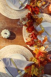 Autumn tablescape on a rustic wood table with woven round placemats, white plates, gray napkins and gold forks, wine glasses, a small pale pumpkin, and a vibrant orange maple-leaf garland centerpiece.