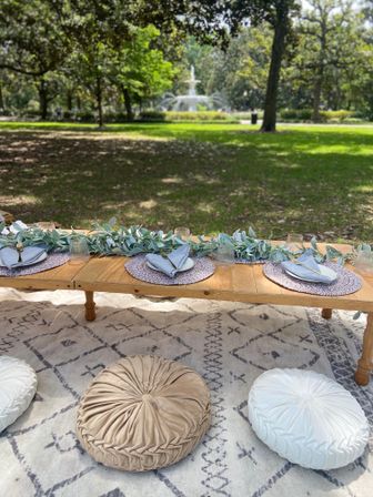 Boho outdoor picnic setup: low wooden table with eucalyptus garland, woven placemats, folded linen napkins, glassware and gold flatware on a patterned rug with round floor cushions in a leafy park with a fountain in the background.