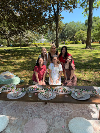 Sunny park picnic with a fountain in the background, six friends posing around a low wooden table set with a floral runner, placemats, glassware and cushions; central guest wearing a white dress and sash.