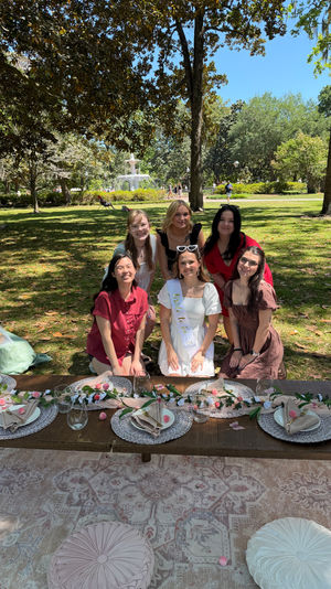 Sunny park picnic with a fountain in the background, six friends posing around a low wooden table set with a floral runner, placemats, glassware and cushions; central guest wearing a white dress and sash.