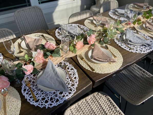 Sunlit backyard patio dining table set al fresco with woven placemats, linen napkins, gold flatware, champagne flutes and a pink rose garland centerpiece