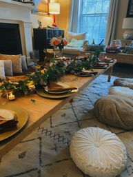 Cozy living room dinner setup with a low wooden table, gold chargers, folded napkins, glassware, greenery garland and tea lights, surrounded by plush round floor cushions, armchair and lamp by a window in a warm boho-chic home.