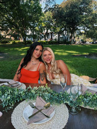 Two friends relaxing at a sunlit park picnic, seated at a rustic table with woven placemats, gold flatware, wine glasses and a leafy eucalyptus garland — summer outdoor dining vibe.