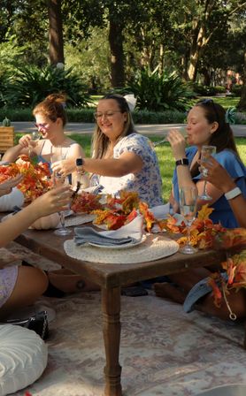 Group of friends toasting at a sunlit outdoor fall picnic in a park, seated around a low wooden table decorated with autumn leaf garlands, small white pumpkins, woven placemats and champagne flutes.