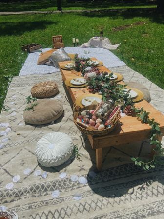 Boho outdoor picnic on a sunny grassy park lawn with a low wooden table, gold chargers, floral garland, round floor cushions and a basket of pastries.