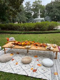 Cozy autumn picnic tablescape on a patterned rug in a city park — low wooden table with gold plates, fall leaf garland and cushions, white ornamental fountain and trees in the background.