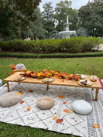 Cozy autumn picnic tablescape on a patterned rug in a city park — low wooden table with gold plates, fall leaf garland and cushions, white ornamental fountain and trees in the background.