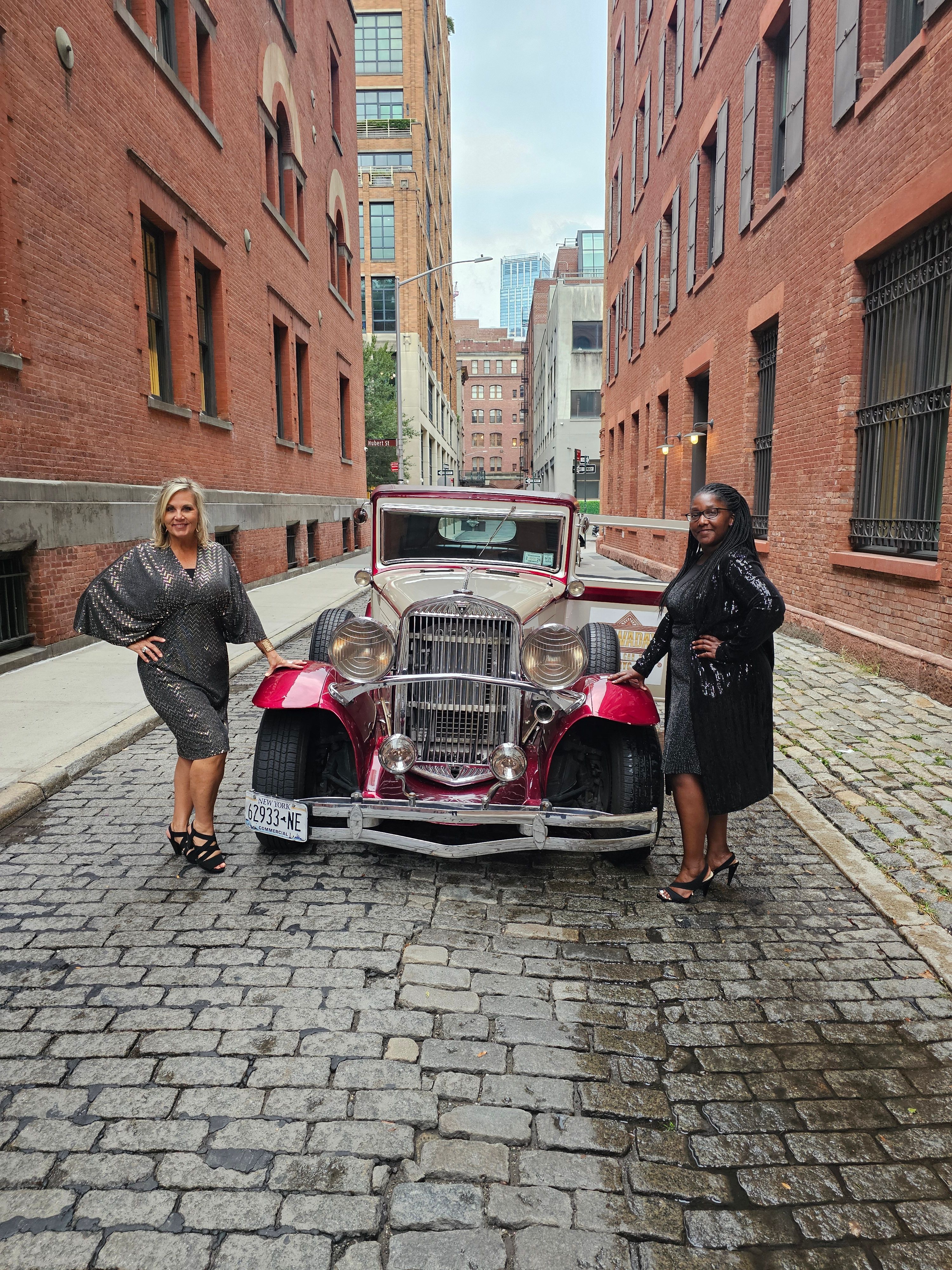 Two women in black sequined dresses pose beside a maroon vintage convertible parked on a cobblestone alley between red brick warehouse buildings with modern city towers visible in the background