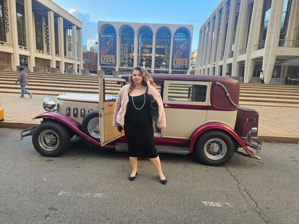 Woman in black dress, pearls and pink faux-fur jacket posing by a cream-and-maroon vintage car with open door on a downtown performing-arts plaza