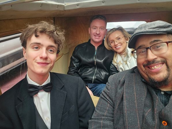 Group selfie of four smiling friends inside a vintage car interior — young man in a tuxedo with bow tie at left, man in a cap and glasses taking the photo at right, couple in back with a leather jacket and light scarf, warm beige upholstery.