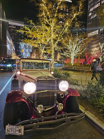 Vintage maroon classic car parked on a New York City street at night, headlights glowing beneath trees and skyscrapers wrapped in warm holiday lights