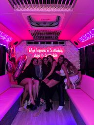 Six friends smiling inside a pink-lit party bus in Scottsdale, posing on bench seats in front of a floral wall and neon sign reading "What happens in Scottsdale, stays in Scottsdale", with metallic balloons and scattered high heels on the floor.
