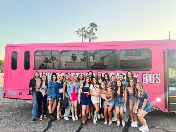 Cheerful group of young women in casual party outfits posing and smiling in front of a bright pink party bus in a parking lot at sunset, palm trees reflected in the bus windows.