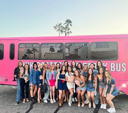 Cheerful group of young women in casual party outfits posing and smiling in front of a bright pink party bus in a parking lot at sunset, palm trees reflected in the bus windows.