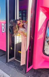 Woman in pink dress and sunglasses leaning out of a bright pink party bus doorway on a sunny street