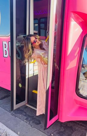 Woman in pink dress and sunglasses leaning out of a bright pink party bus doorway on a sunny street