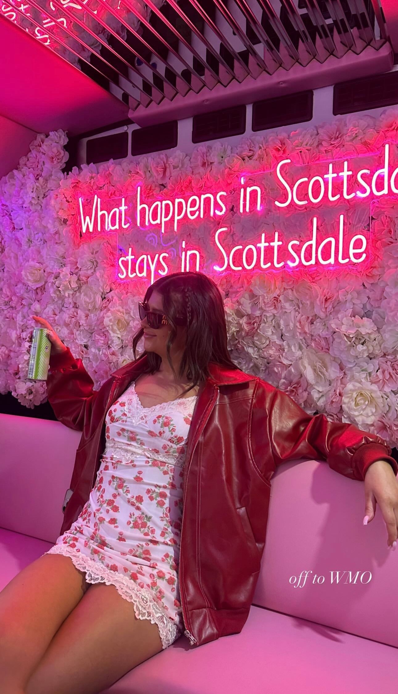 Person in red leather jacket and floral mini dress lounging on a pink couch under a bright pink neon sign reading What happens in Scottsdale stays in Scottsdale, floral wall backdrop, holding a canned drink