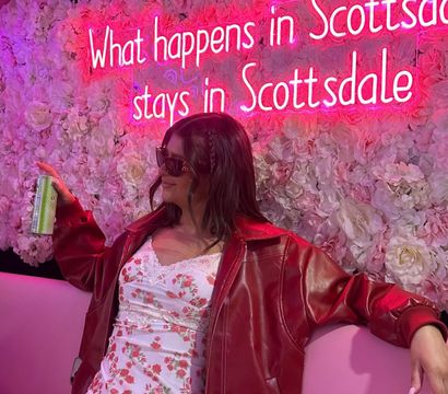 Person in red leather jacket and floral mini dress lounging on a pink couch under a bright pink neon sign reading What happens in Scottsdale stays in Scottsdale, floral wall backdrop, holding a canned drink