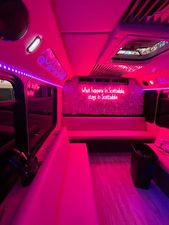 Vibrant pink-lit party bus interior with wraparound bench seating, LED accent strips, floral wall and neon sign reading "What happens in Scottsdale, stays in Scottsdale".