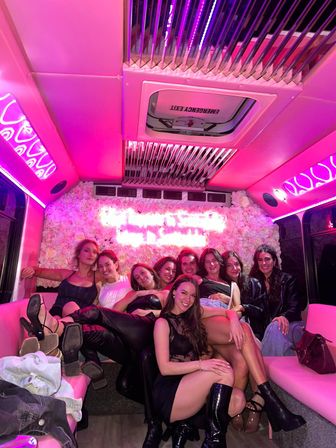 Group of women laughing and posing on a pink‑lit party bus with a floral wall and neon sign, lounge seating, high heels and boots conveying a fun night‑out celebration.