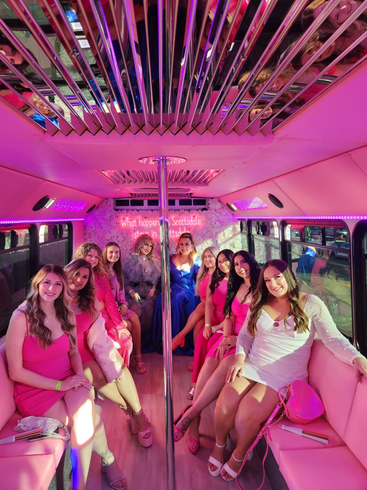 Group of women in pink and white dresses smiling and celebrating inside a neon-pink party bus with a mirrored ceiling, center dance pole, and floral, neon-lit backdrop.