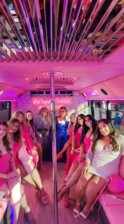 Group of friends in pink and white dresses inside a pink party bus in Scottsdale, mirrored ceiling and chrome pole, neon pink lights and a floral wall creating a bachelorette-party vibe.