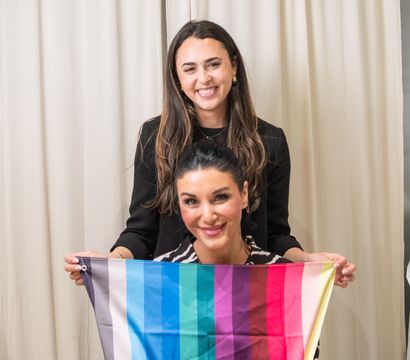 Indoor portrait of two smiling women against cream curtains, the woman in front holding a bright multicolor vertical-striped scarf in shades of gray, blue, teal, purple and red.