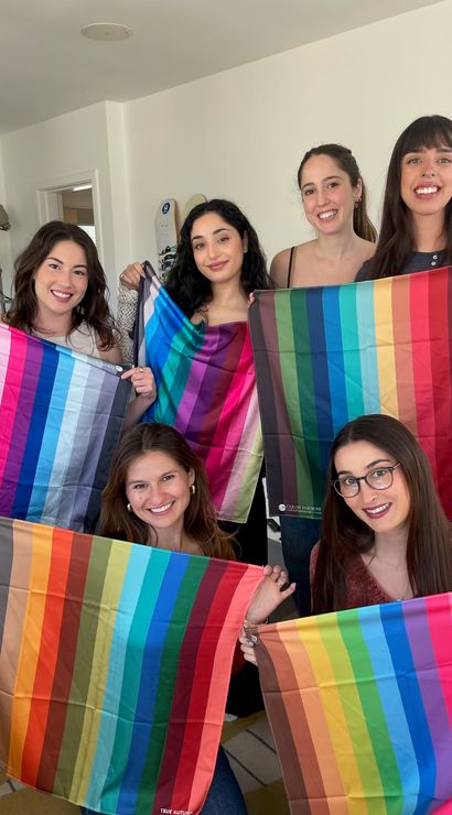 Six smiling women in a bright living room holding vibrant multicolored striped fabric swatches and color-palette scarves, posing for a playful group photo.