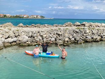 Three people cheering in shallow turquoise water of a tropical coastal cove, using a colorful paddleboard as a floating drinks station near a rocky breakwater with resort homes on the distant shoreline.
